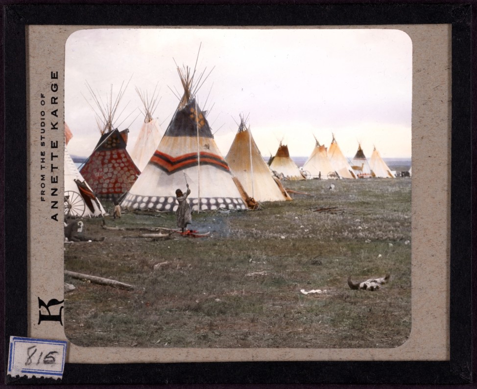 Woman chopping firewood, Eagle tipi in foreground, Star tipi on left. Hand-painted lantern slide by photographer Walter McClintock (1870-1949) of the Blackfoot Indians of Montana. (Yale Collection of Western Americana, Beinecke Rare Book and Manuscript Library)