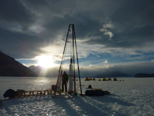 Ice core driller Tanner Kuhl with the blue ice drill on Taylor Glacier in Antarctica. The field camp is visible in the background. (© Xavier Fain/Oregon State University)